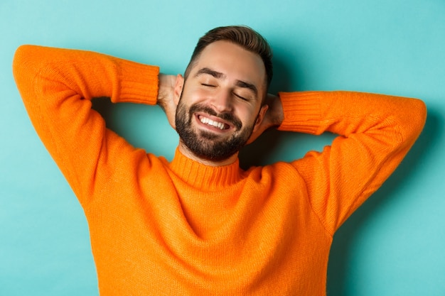 Carefree young man daydreaming, resting with hands behind head and closed eyes, relaxing over turquoise background in orange sweater.