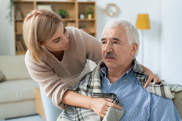 Careful and affectionate daughter taking care of her sick senior father while wrapping him in plaid at home