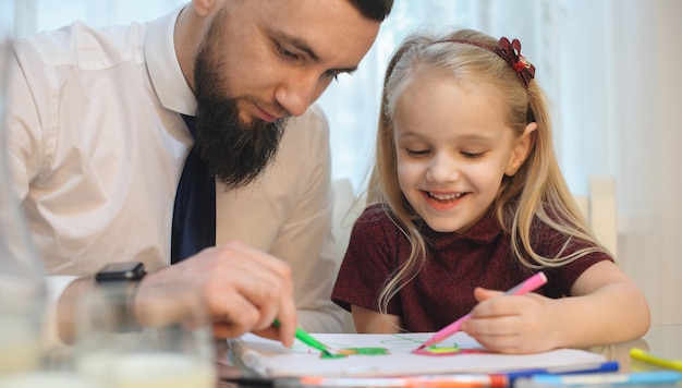 Careful caucasian businessman drawing with his small girl sitting at the table dressed in a white shirt with tie