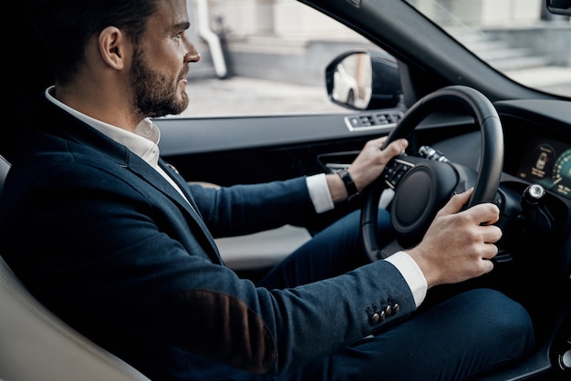 Careful driving. Handsome young man in full suit looking straight and smiling while driving a car