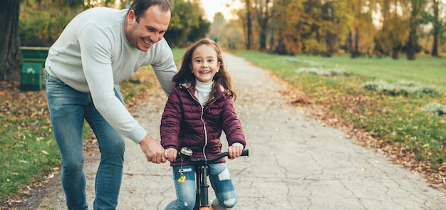 Careful father teaching his daughter how to ride the bike helping her in the park smiling