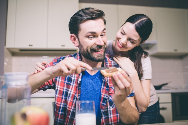 Careful girl stands behind guy and loks at him. She smiles. Man puts some chocolate paste on toast and look at her. He smiles as well. They are happy.