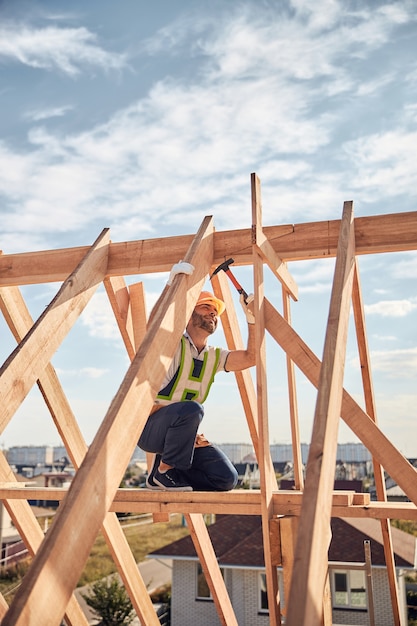 Careful man wearing a hardhat and holding a hammer while kneeling on a narrow wooden bar up high