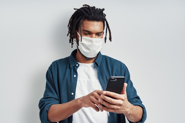 의료 얼굴 마스크를 착용하고 회색 벽에 서있는 동안 스마트 폰을 사용하는 조심스러운 젊은 남자