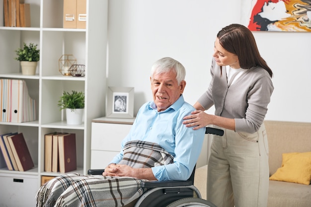 Заботливая молодая медсестра разговаривает с пожилым мужчиной в инвалидной коляске, навещая его дома