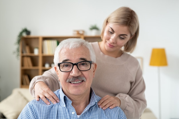 Attenta giovane donna che fa massaggio delle spalle a suo padre senior che guarda l'obbiettivo con il sorriso in ambiente domestico