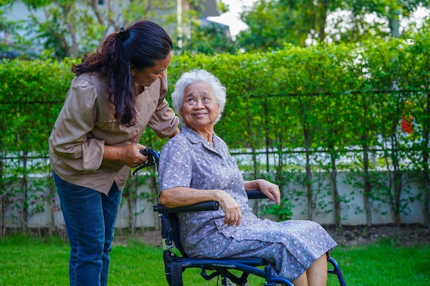 간병인은 공원 의료 개념에서 휠체어에 앉아 있는 아시아 노인 여성 장애 환자를 돕습니다.