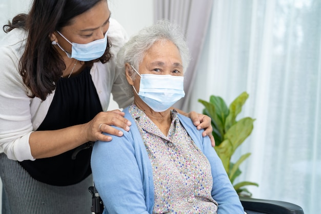 간병인은 안전 감염 코비드-19 코로나바이러스를 보호하기 위해 휠체어에 앉아 안면 마스크를 쓴 아시아 노인 또는 노년 여성을 돕습니다.
