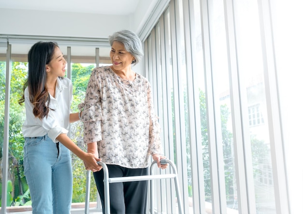 Caregiver smiling young asian female daughter or grandchild supporting help elderly woman mother or grandparents try to walk with walker frame at home on green nature background senior healthcare