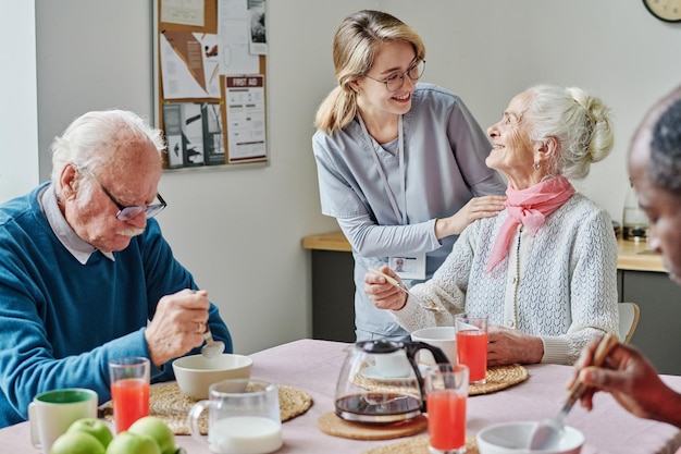 Caregiver talking to senior people and helping them while they eating at table