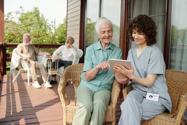 Caregiver in uniform showing something to senior woman on tablet screen