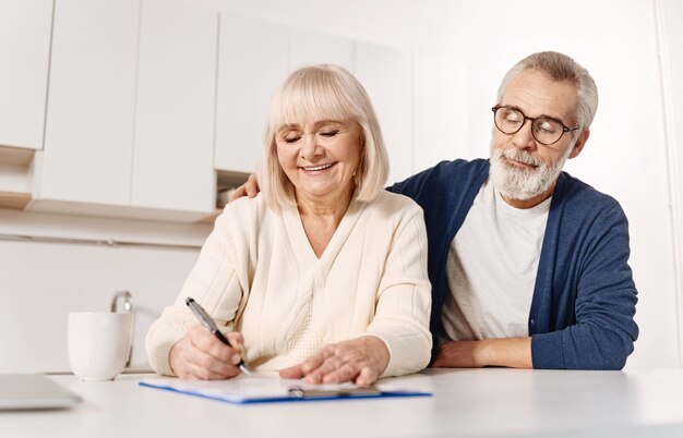 Foto prendersi cura del futuro dei nostri nipoti. donna anziana positiva armonica sorridente che si siede a casa accanto a suo marito ed esprime la felicità mentre firma i documenti