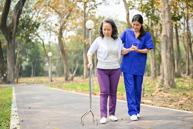 돌보는 아시아 여성 간병인이 공원에서 지팡이를 짚고 걷는 노부인을 돕고 있습니다.
