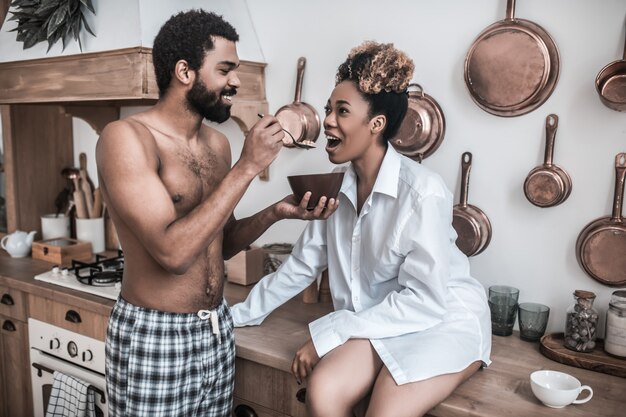 Caring attitude. Caring young bearded happy man standing with plate and spoon near wife sitting with open mouth in kitchen