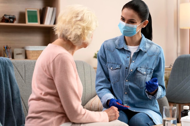 Caring brunette nurse talking with senior woman at home Young practitioner wearing protective mask taking care of the elderly woman