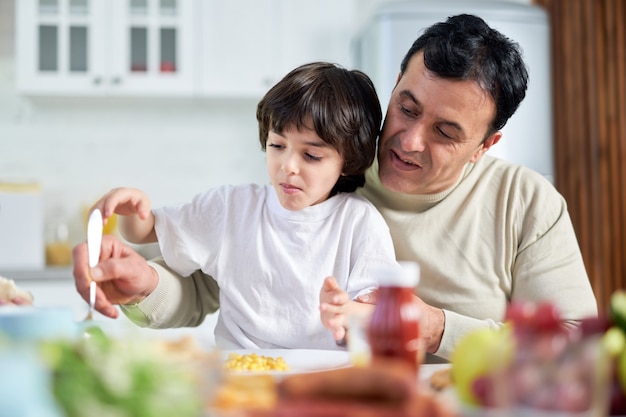 Caring dad smiling middle aged latin father serving his son sitting with him at kitchen