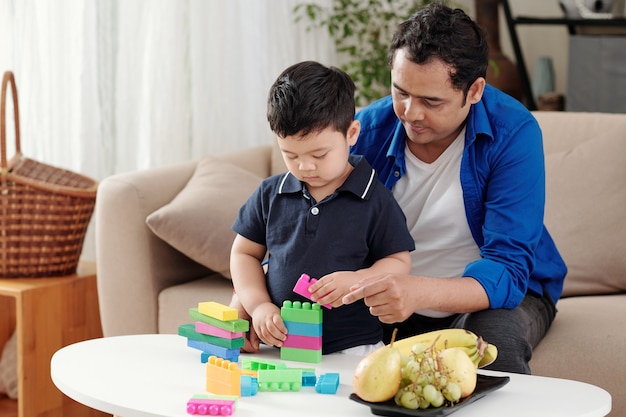 思いやりのある父親は、幼い息子が色とりどりの木製とプラスチックのブロックから塔を建てるのを手伝っています