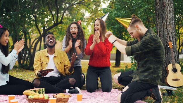 Caring friends are bringing cake to African American man sitting on blanket in park on picnic with closed eyes he is blowing candles and laughing enjoying surprise