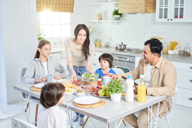 Caring hispanic woman serving salad for her husband and children, standing in the kitchen. Latin family having dinner together at home. Selective focus