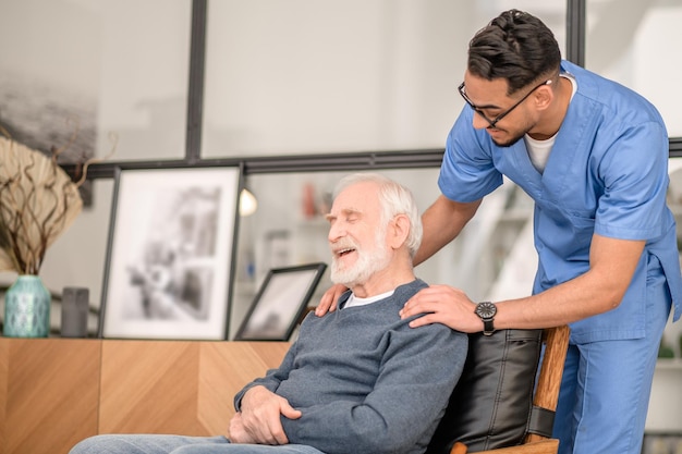 Caring in-home nurse in uniform leaning over a happy old man seated in the armchair