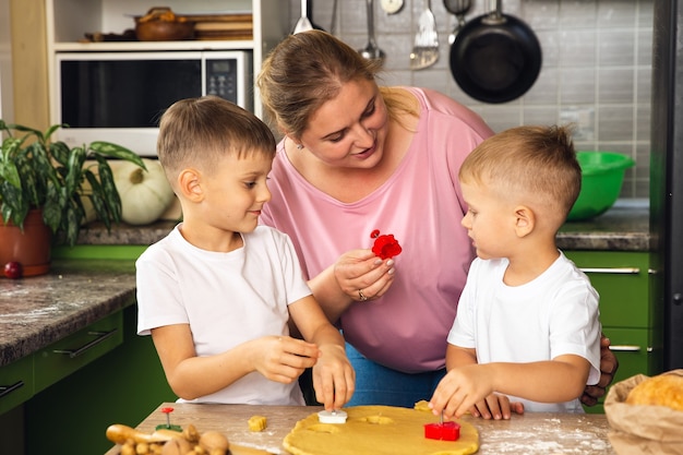 La madre premurosa aiuta i figli piccoli in età prescolare a preparare i biscotti, la mamma amorevole sorridente impara a cucinare con i bambini piccoli, a preparare il pranzo nel fine settimana in cucina insieme