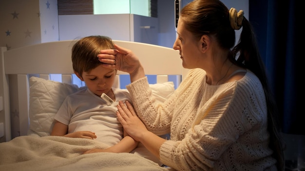 Caring mother measuring temperature to her sick little son lying in bed at night Children illness