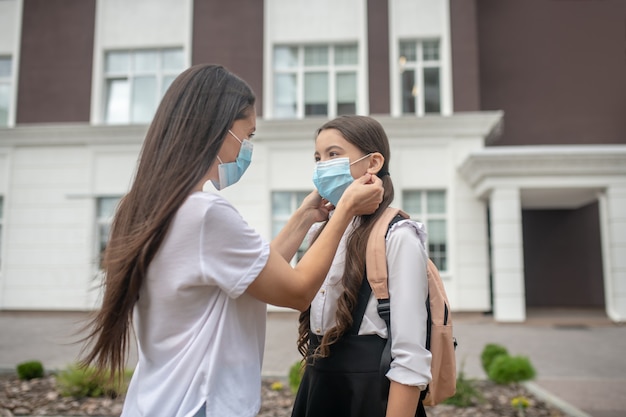 Madre premurosa che indossa la maschera protettiva per sua figlia studentessa in piedi nel cortile della scuola