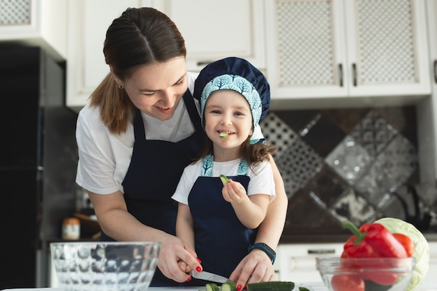 Madre premurosa che insegna alla piccola figlia a cucinare insalata in cucina, giovane mamma e adorabile bambina carina che indossa un grembiule tagliando le verdure con un coltello sul bancone, in piedi in cucina a casa