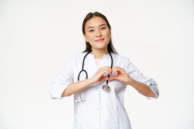 Caring nurse in hospital or clinic wears uniform, shows heart sign, taking care of patients, stands over white background