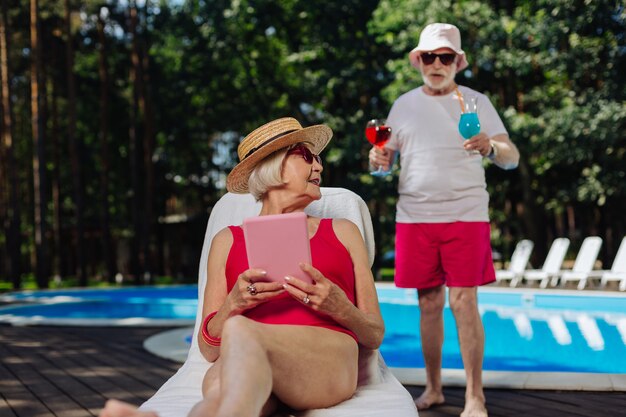 Uomo in pensione premuroso che porta alcuni cocktail estivi per la moglie che prende il sole vicino alla piscina