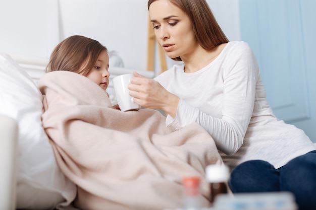 Photo caring woman helping her daughter to recover and drinking tea while sitting on the couch