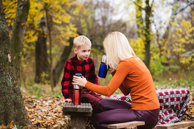 Caring woman try hot drink from stainless thermos before give it to little preteen boy. Happy cozy family picnic at wooden bench of autumn park