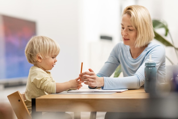 Caring young caucasian mother and small son drawing painting in notebook at home together loving mom