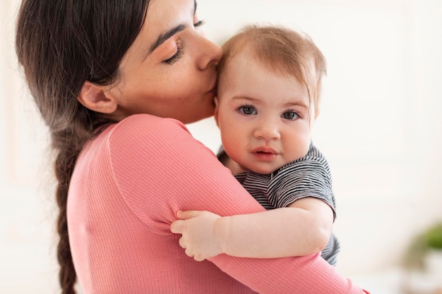 Заботливая молодая мать держит и целует милую маленькую девочку, мама и очаровательный ребенок, сближающиеся дома, копируя пространство