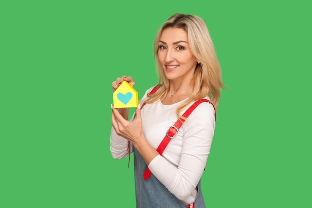 Caring for your home, real estate insurance. Portrait of positive beautiful adult blond woman in urban style overalls holding paper house with heart inside. studio shot isolated on green background