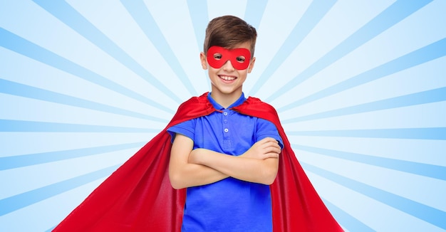 carnaval, kindertijd, macht, gebaar en mensenconcept - gelukkige jongen in rode superheldencape en masker over blauwe burst-stralenachtergrond