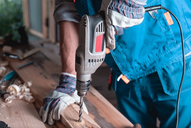 Carpenter in overalls and gloves drills a wooden board in the home workshop