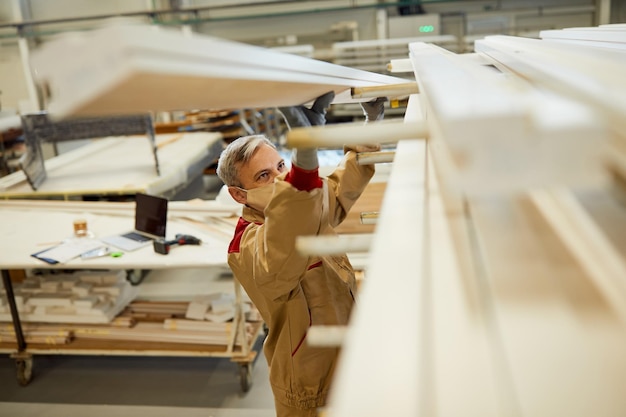 Carpenter with face mask while taking wood planks from a shelf in a workshop