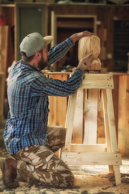 Плотник в рабочей одежде пила, чтобы вырезать скульптуру из деревянной головы человека в мастерской