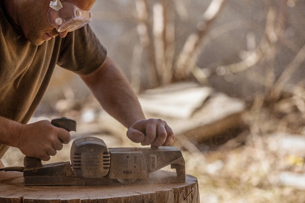 Carpenter working with electric planer on wooden stump in the open air, wearing goggles.