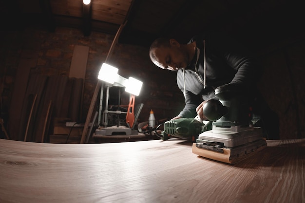 A carpenter works in a workshop Joiner's grinders furniture manufacturing A carpenter is grinding a wooden part with an electric sander