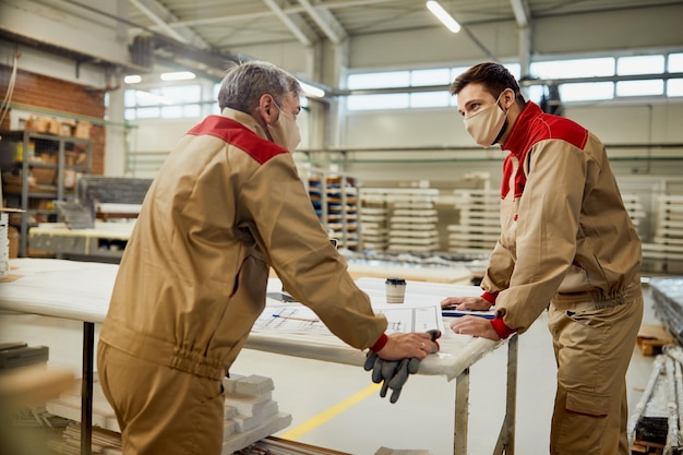 Carpenters with face masks talking while examining blueprints in a workshop