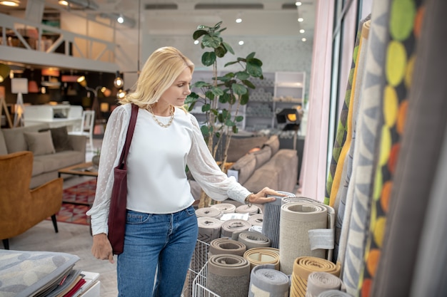 Carpets and rugs. Serious woman in a furniture store standing opposite the carpets with interest looking, hand on the rug.