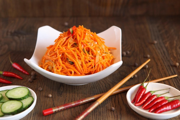 Carrot Korean vegetable salad in a bowl with chopsticks on wooden oak table and green salad leafs, fresh red chilli peppers. Spicy Korean-style carrot salad on a dark background. Koryo-saram cuisine.