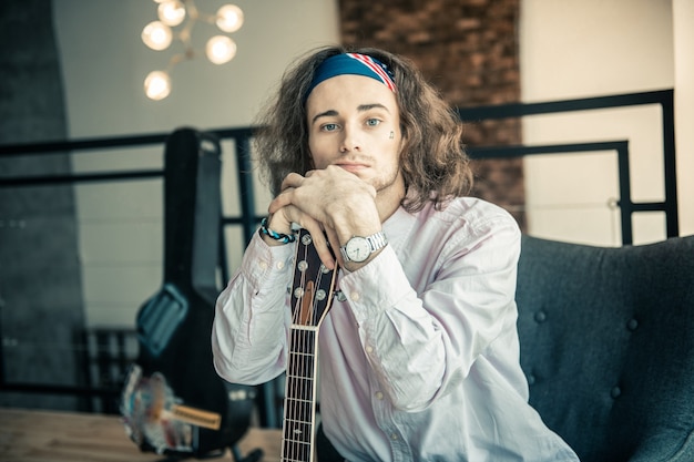 Carrying acoustic guitar. Tired handsome man with tattoo on the face putting both hands on the neck of his guitar