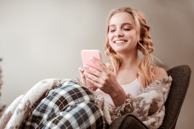 Carrying mobile phone. Joyful appealing lady being covered in wool blanket while resting in armchair