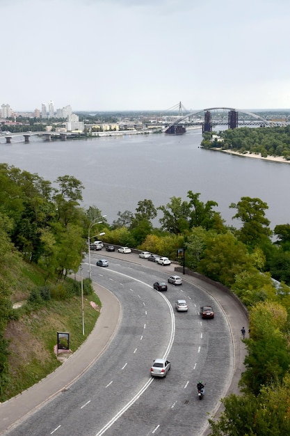 Cars drive along the winding asphalt highway In the background is a wide river and a bridge