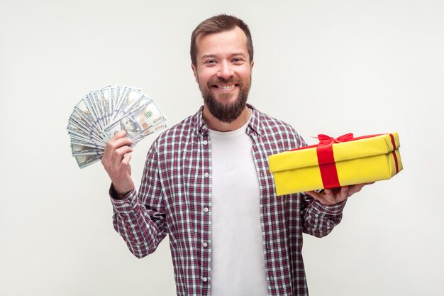 Cashback and present for buyer Portrait of happy bearded man in casual plaid shirt holding wrapped box and dollars smiling satisfied with gift indoor studio shot isolated on white background