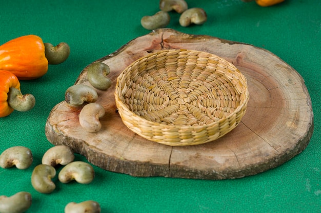 Яблоко кешью и семена кешью расположены на зеленом текстурированном фоне с деревянным куском и маленькой корзиной, расположенной рядом с селективным фокусом