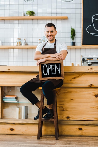 Cashier in apron sitting on chair near wooden bar counter and holding chalkboard with open lettering